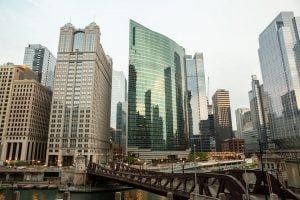 Photo of five large office buildings in Chicago with the skyline reflected in the glass of the center building, on a cloudy day