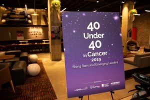 Photo inside dimly lit conference room, with purple poster stating "40 under 40 in Cancer" in foreground