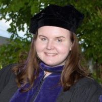 A young woman with light skin and warm brown hair smiles at the camera, wearing a cap and gown.