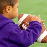 Photo of past Super Fest event, child with short black hair sitting on football field, holding football in both hands. Child's face is in profile, slightly faced away from camera.