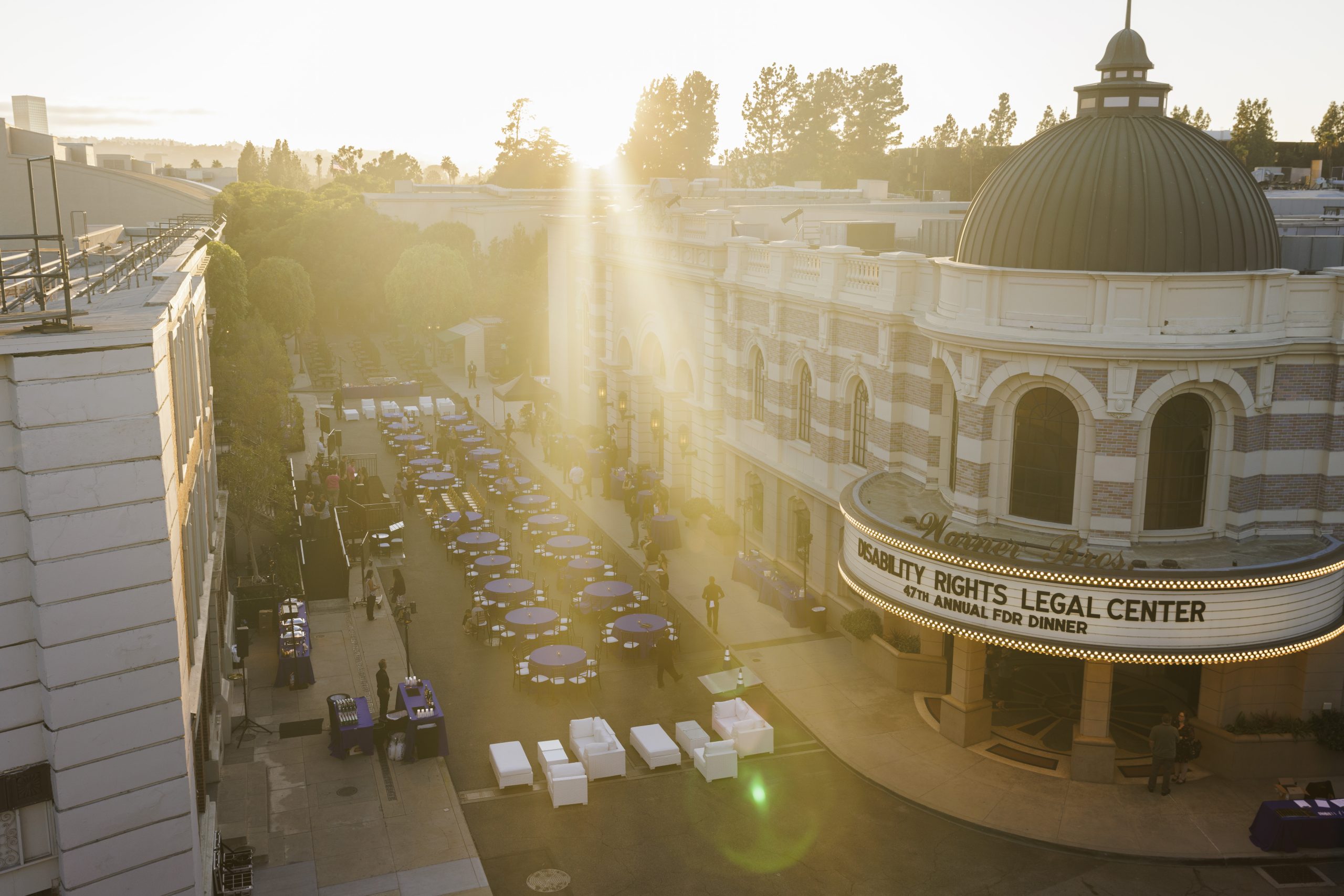 A panned-out image of Brownstone Street set with a lens flare from the setting sun in the distance. Guests haven't arrived yet; rows of tables are covered in royal blue tablecloths; a marquee reading "Disability Rights Legal Center - 47th Annual FDR Dinner" is visible on the front of a rotunda on the end of a theater (not visible).