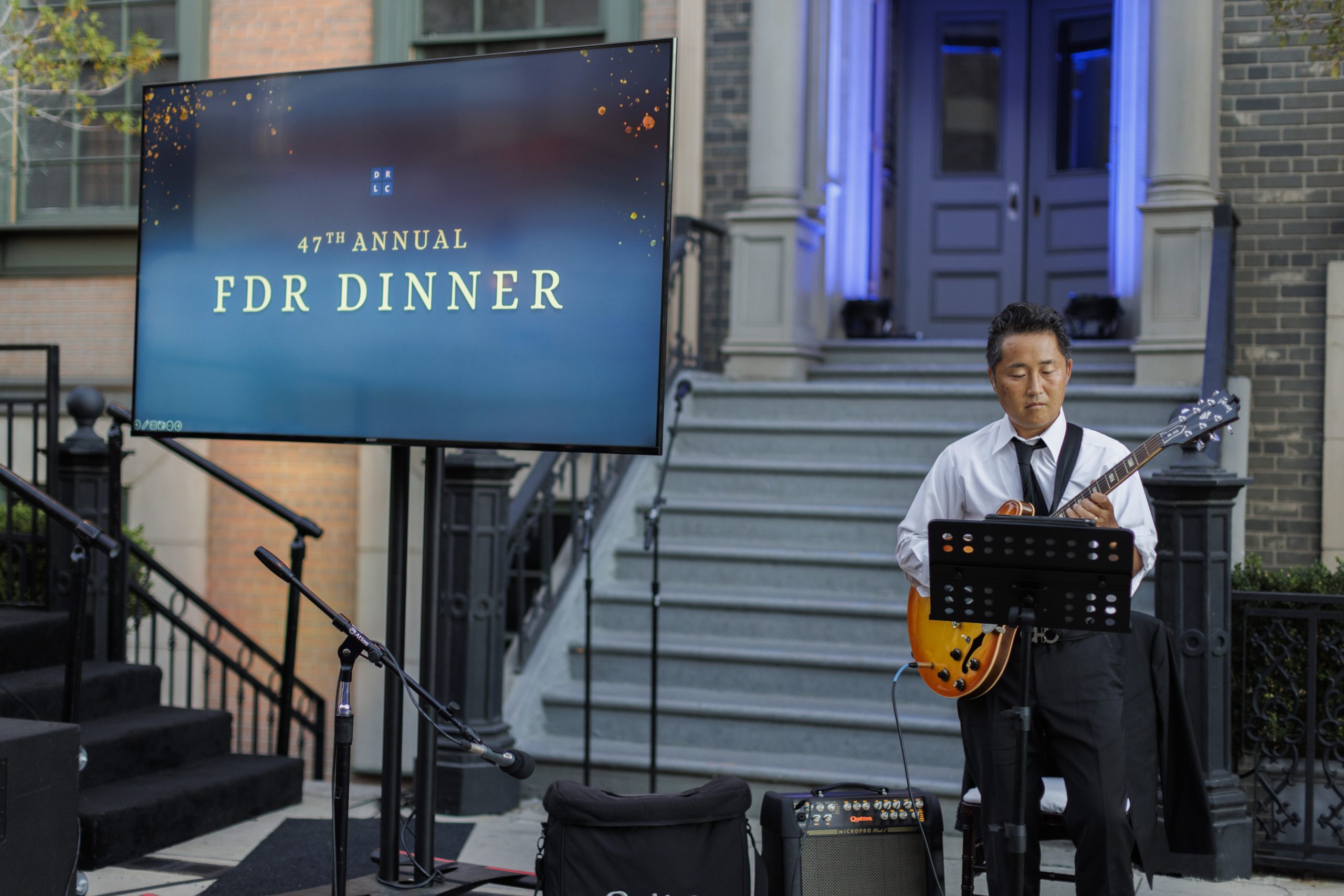 Candid shot of musician Gaku, playing guitar and standing to the left of a large monitor that reads "47th Annual FDR Dinner" with gold text on a blue background. Behind Gaku is a New York-style front stoop.