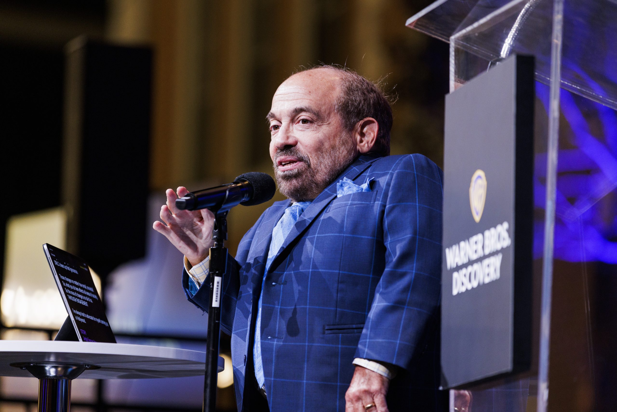 Danny Woodburn, wearing a navy blue suit, addresses the crowd from the microphone and gestures with his right hand. To his right is a clear podium with a sign bearing the words "Warner Bros." on the front.