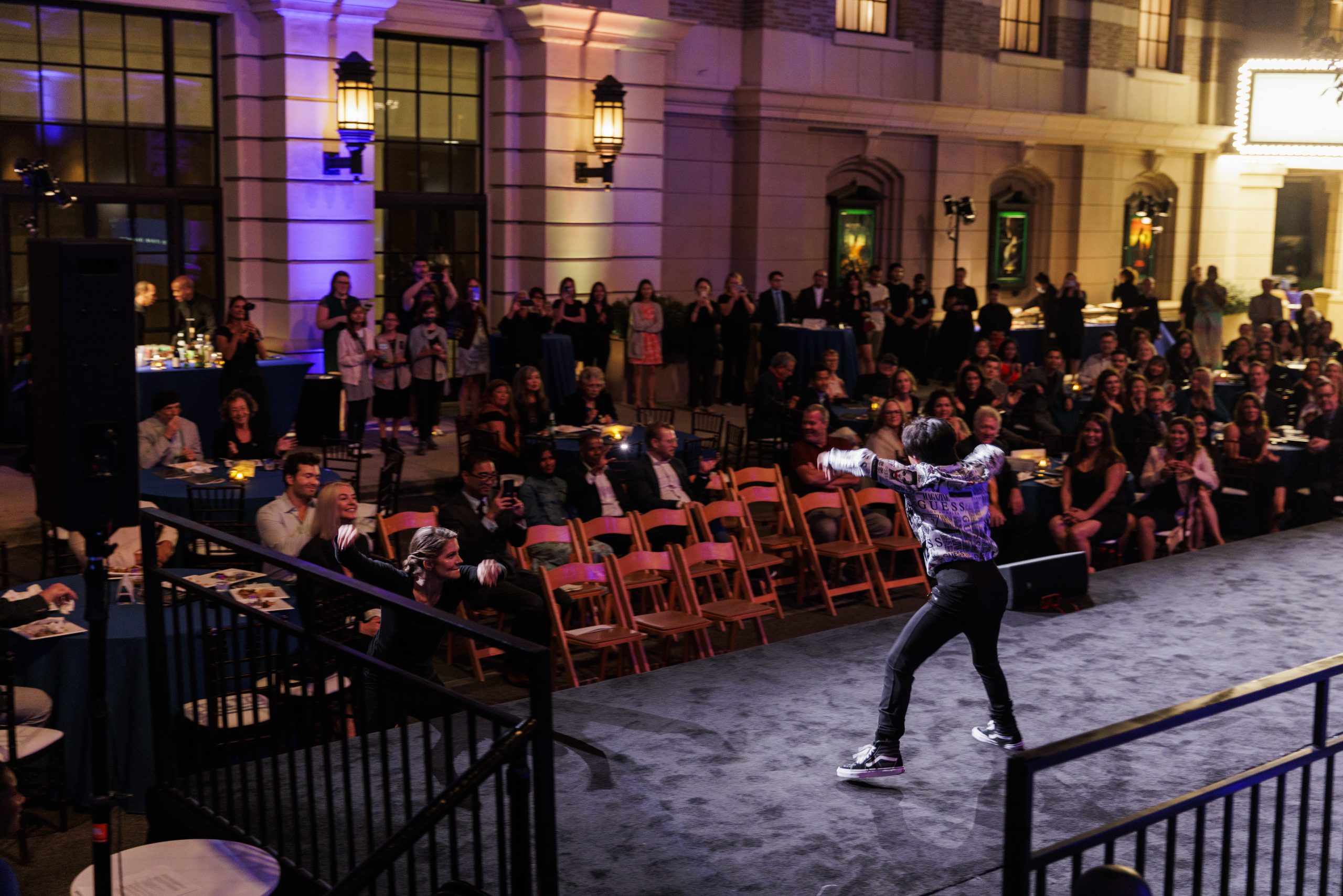 From behind the stage, a shot of a dancer with short black hair, arms outstretched, legs in a wide lunge. The crowd beyond him is dark but visible, clapping to the music.