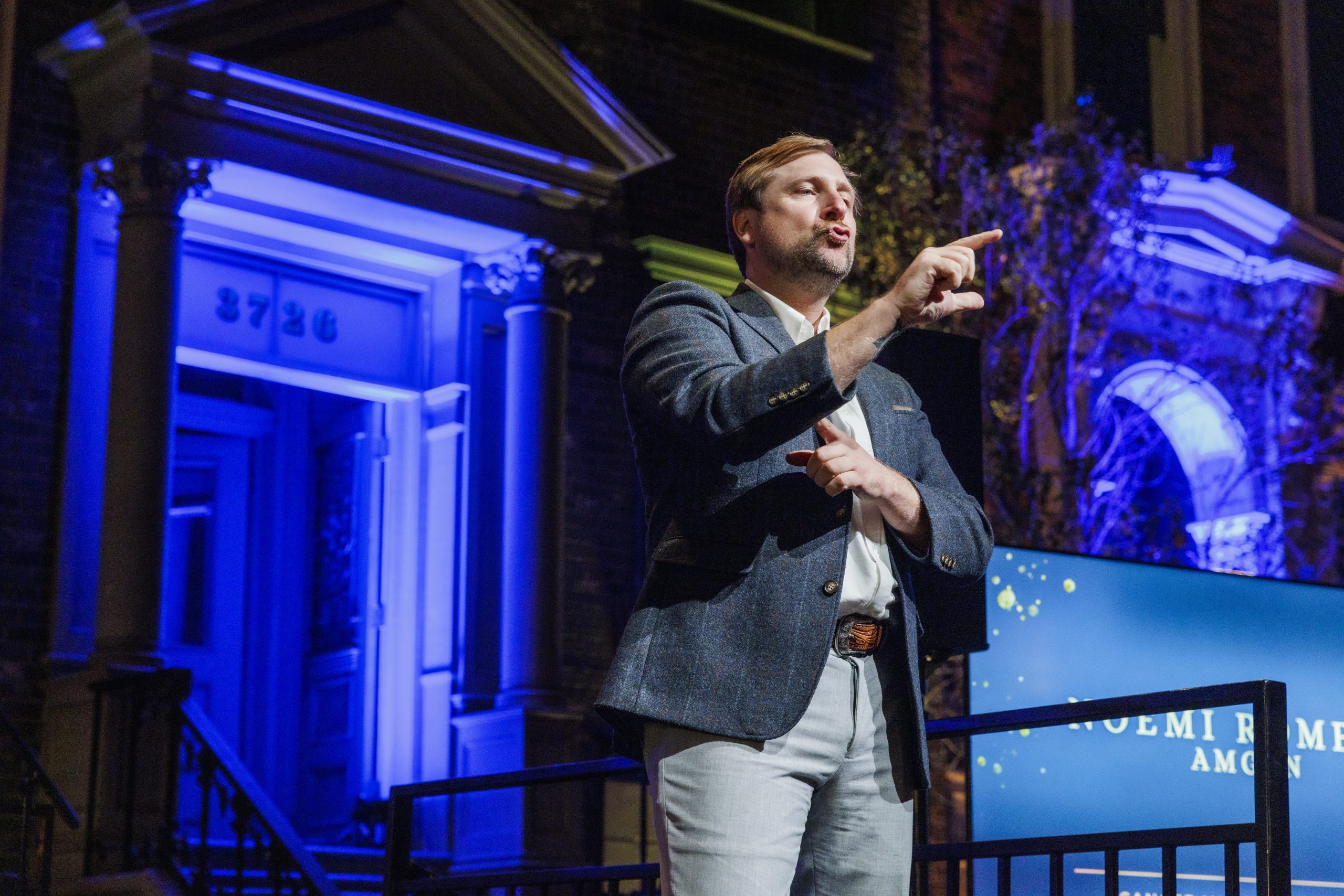 ASL interpreter Justin Maurer, with short blond hair, wearing a dark suit jacket and light pants, interprets on stage, one arm outstretched in front of him, pointing, the other across his chest