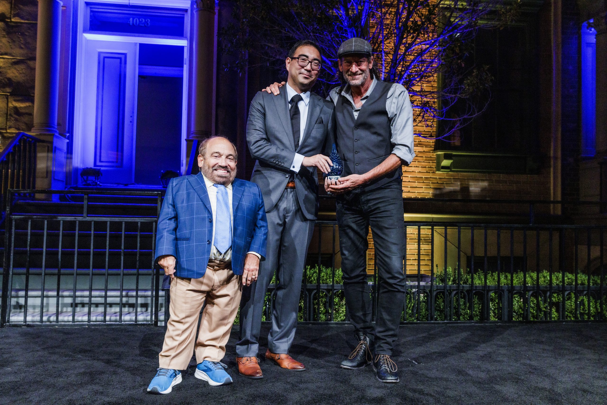 Danny Woodburn, Jordan Kough, and Troy Kotsur pose for a photo with Kotsur's DREAM award, a blue glass teardrop, on stage
