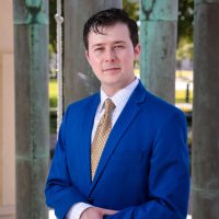 Andrew Gray wears a blue suit, white shirt, and gold tie. He stands, with hands folded, looking at the camera. Behind him are four large pillars, out of focus.