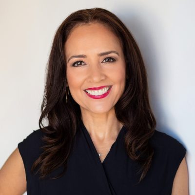 Sylvia Torres-Guillen, wearing a solid black top, long wavy dark hair, smiles at the camera for a head shot