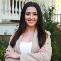 Ashly Villa Ortega wears long dark hair, white shirt, with tan blazer, folds her arms, and smiles for a picture; behind her is a white porch with columns and greenery.