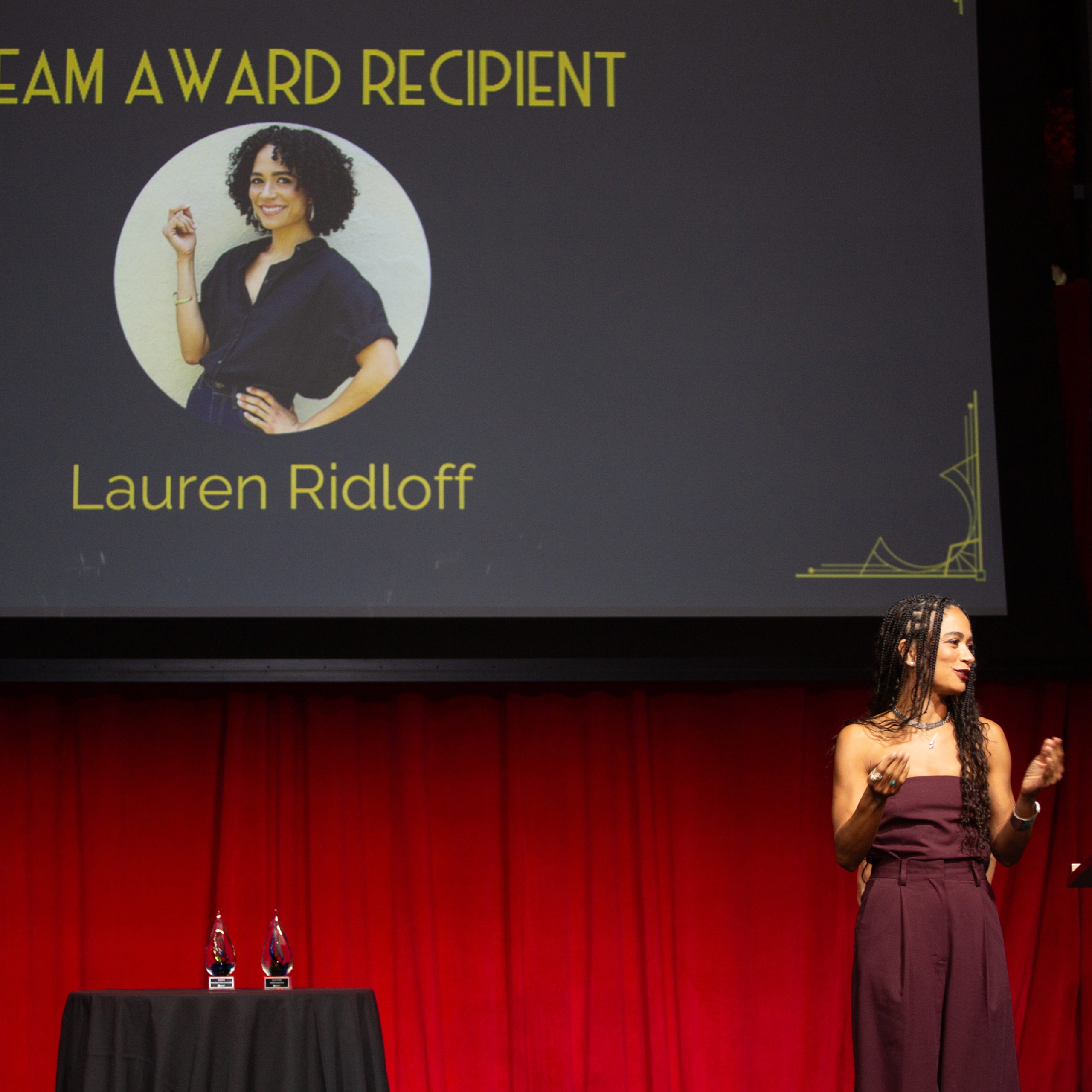 Lauren Ridloff, wearing long braids and a burgundy dress, signs her speech in ASL from the stage. Behind her the projector screen reads "DREAM Award: Lauren Ridloff"