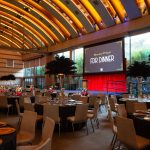 Staged Guerin Pavilion at Herscher Hall at Skirball Cultural Center. Arched wooden ceilings over tan carpet, blue stage with red back drop, very tall black feather centerpieces over round tables with black table cloths. Garden is visible through back wall with floor to ceiling windows.