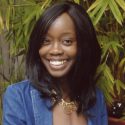 Shika Pappoe wears her dark hair down and wavy, with a gold necklace, white shirt, and blue blazer.